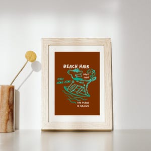 May include: Framed art print with a brown background and a teal illustration of a creature surfing. The text reads "BEACH HAIR DON'T CARE" and "THE OCEAN IS CALLING". The frame is a light wood color.