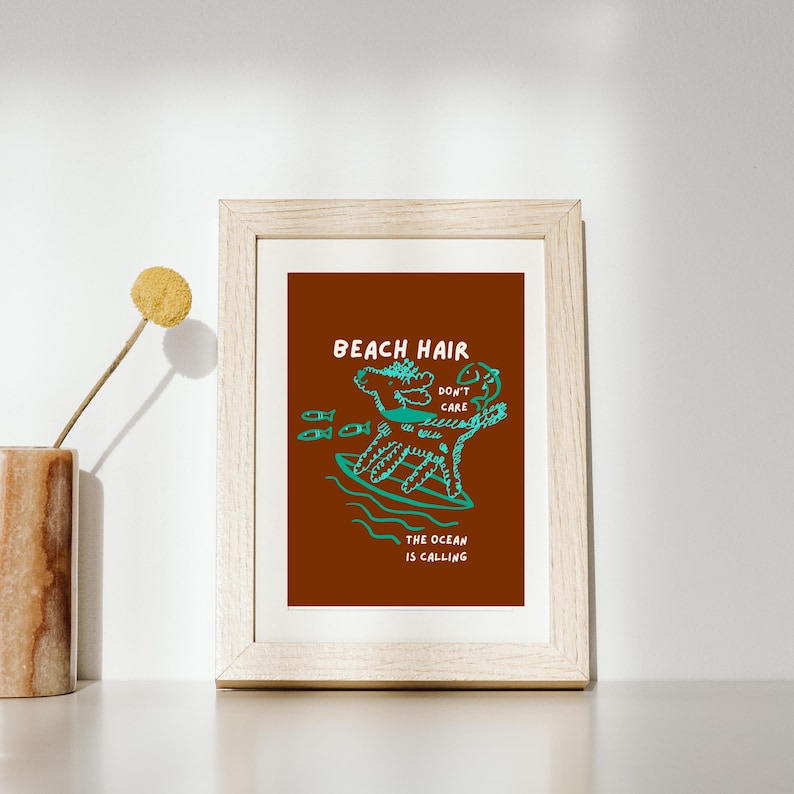 May include: Framed art print with a brown background and a teal illustration of a creature surfing. The text reads "BEACH HAIR DON'T CARE" and "THE OCEAN IS CALLING". The frame is a light wood color.