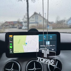 May include: Interior car shot with a digital display showing a map and music interface. A clear TOFAS logo car air freshener hangs from the rearview mirror. The car's air vents are visible below the display.