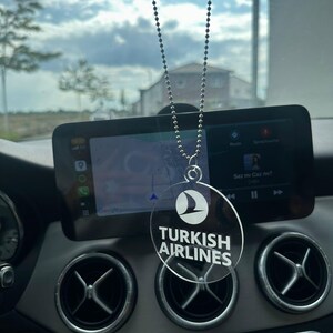 Könnte beinhalten: Ein klarer, kreisförmiger Auto-Lufterfrischer mit dem Logo und Text von Turkish Airlines. Der Lufterfrischer hängt an einer silbernen Perlenkette, sichtbar vor einem Armaturenbrett und einem bewölkten Himmel.