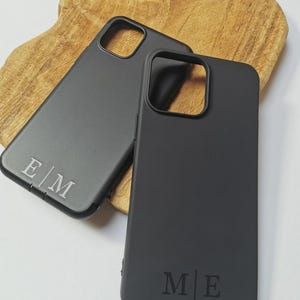 Könnte beinhalten: Zwei schwarze Handyhüllen mit personalisierten Initialen. Eine Hülle zeigt "E|M" und die andere "M|E". Die Hüllen liegen auf einem Holzuntergrund und zeigen ein minimalistisches Design.