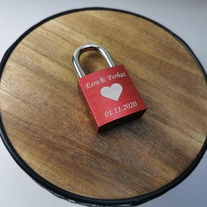 Può includere: Un lucchetto rosso con un grillo argentato, inciso con i nomi "Esra & Ferhat", un cuore bianco e la data "01.11.2020". Il lucchetto è posto su una superficie rotonda in legno con bordo scuro. Un regalo personalizzato per matrimonio o anniversario.