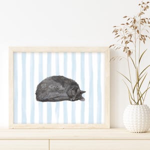 May include: A framed print featuring a watercolor illustration of a sleeping gray cat. The artwork is set against a light blue and white striped background. The frame is light wood. A white vase with decorative stems is to the right.