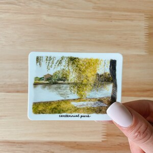 Pu&ograve; includere: Un'illustrazione ad acquerello di una scena di parco con un lago, alberi e un piccolo edificio in lontananza. Il testo "centennial park" &egrave; scritto in basso nell'immagine.