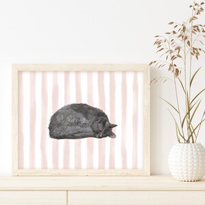 May include: A framed print featuring a watercolor illustration of a sleeping gray cat. The background has vertical pink and white stripes. The artwork is displayed on a light-colored wooden surface, with a white vase of dried plants to the right.