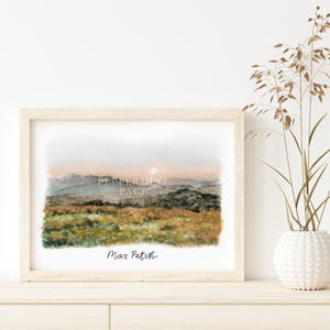 May include: A watercolor painting of a landscape with mountains in the background and a field of grass in the foreground. The sky is a soft pink and orange, and the sun is setting behind the mountains. The painting is framed in a light wood frame and has the text "Max Patch" written in black ink at the bottom.