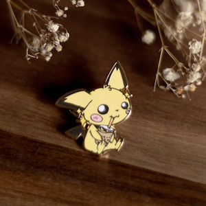 Könnte beinhalten: Ein gelber Pikachu-Emaille-Pin mit einem Getränk, auf einer Holzoberfläche. Der Pin hat schwarze und weiße Details mit einem lächelnden Gesichtsausdruck. Getrocknete Blumen und Blätter im Hintergrund schaffen eine weiche, natürliche Ästhetik.
