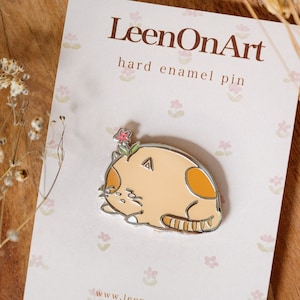 Puede incluir: Un pin de esmalte duro con un gato de dibujos animados con una flor en la cabeza. El gato está enroscado y tiene un esquema de color naranja claro y blanco. El pin está en una tarjeta blanca con el texto "LeenOnArt hard enamel pin" y "www.leenonart.com".