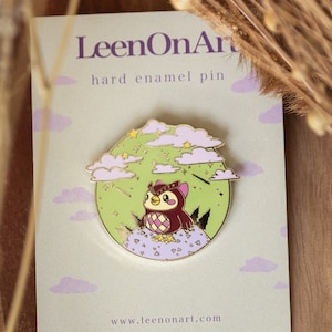 Könnte beinhalten: Eine goldene Emaille-Anstecknadel mit einem Cartoon-Eulenmotiv, das auf einem Hügel mit lila Blumen und Wolken im Hintergrund sitzt. Die Anstecknadel trägt den Schriftzug "LeenOnArt" und "hard enamel pin" auf der Karte, auf der sie liegt. Die Karte enthält auch die Website "www.leenonart.com".