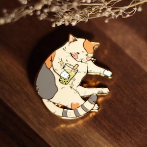 Puede incluir: Un pin de esmalte dorado con un gato de dibujos animados en crema, naranja y gris, sosteniendo un té de burbujas verde. El pin está sobre una superficie de madera oscura. Un accesorio con temática de gatos.