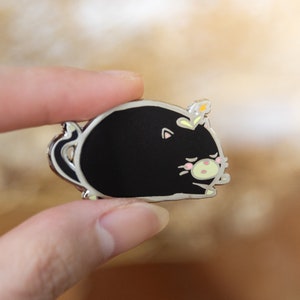 Puede incluir: Un pin de esmalte negro de un gato de dibujos animados con una corona de flores. El gato está enroscado y tiene la nariz rosa y bigotes blancos.