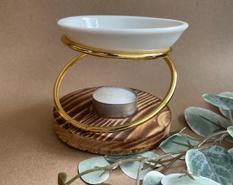 May include: A white ceramic oil burner with a gold metal ring, sitting on a wooden base with a tea light candle in the center.
