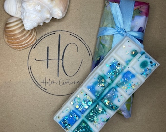 Könnte beinhalten: Ein rechteckiger Wachsschmelzriegel mit blauen und türkisfarbenen Akzenten und Glitzer, neben einem mit einem blauen Band gebundenen verpackten Artikel und Muscheln. Das Logo "HC Holme Creations" ist sichtbar.