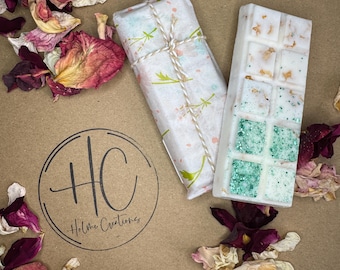 Könnte beinhalten: Zwei Wachsschmelzriegel, einer in florales Papier mit Bindfaden verpackt, der andere weiß mit grünen Glitzerquadraten und getrockneten Blütenblättern. Der Hintergrund ist eine braune Oberfläche mit Blütenblättern und einem Logo.