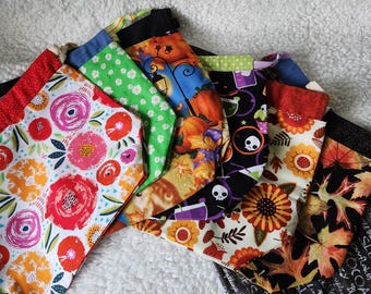 May include: A collection of fabric drawstring bags featuring various patterns including red and orange floral prints, green daisy motifs, orange pumpkins, black skull designs, yellow sunflowers, and autumn leaves with text on a black background.