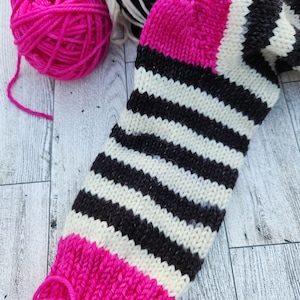 May include: A pink, white, and black striped knitted sock with a pink cuff. The sock is lying on a wooden surface.