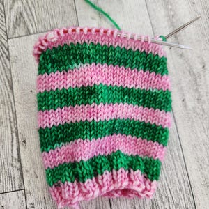 May include: A partially knitted sock in progress, featuring alternating horizontal stripes of green and pink yarn. Knitting needles are inserted into the top row of stitches. A ball of green yarn is visible in the background.
