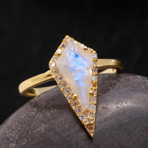 May include: A gold ring featuring a large, clear, iridescent gemstone set in a diamond-shaped setting. The gemstone is surrounded by smaller, clear gemstones.