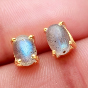 Peut inclure: Une paire de boucles d'oreilles clous dorées avec des pierres précieuses en labradorite de forme ovale. Les pierres précieuses sont de couleur bleu-gris avec des reflets irisés.