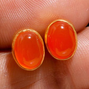 May include: A pair of oval-shaped, orange-red gemstone stud earrings. Each stone is set in a gold-colored bezel. The earrings are displayed against a skin-toned background.