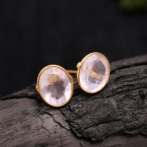 Op de afbeelding: Een paar goudkleurige oorstekers. Elke oorbel heeft een grote, ovale, lichtroze edelsteen gezet in een gouden bezel. De oorbellen worden gepresenteerd op een donker, getextureerd oppervlak, wat een contrast creëert.