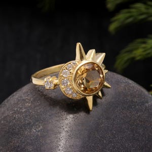 Könnte beinhalten: Goldring mit einem großen, runden, topasfarbenen Edelstein, der von kleineren, klaren Edelsteinen umgeben ist. Der Ring hat ein Sonnenstrahlendesign mit spitzen Strahlen.