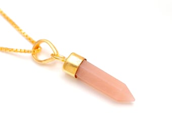 Può includere: Una collana con catena dorata e un ciondolo a punta di cristallo rosa. Il ciondolo è attaccato alla catena con un connettore dorato a forma di cuore.
