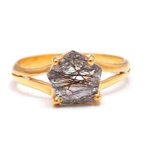 May include: A gold ring featuring a faceted, geometric gemstone with black inclusions. The ring has a split shank design and is set against a white background. The gemstone is approximately 0.75 inches in diameter.