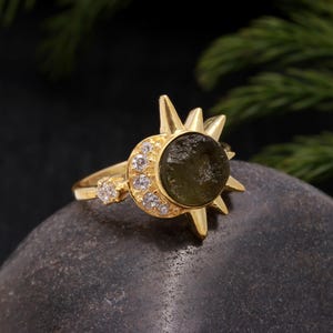 Könnte beinhalten: Goldring mit einem grünen Edelstein, der in einem Sonnenstrahlendesign mit klaren Zirkonia-Steinen um den Umfang herum gefasst ist.