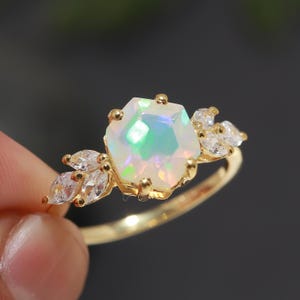 Peut inclure: Une bague en or avec une grande opale irisée sertie dans un hexagone. La bague est ornée de petites pierres précieuses claires de chaque côté de l'opale.