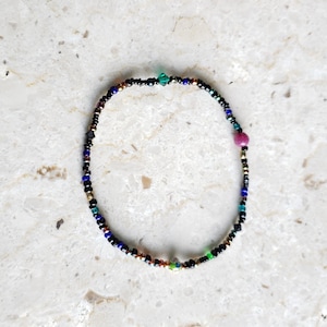 May include: A delicate bracelet made of black beads with accents of blue, green, and gold beads. The bracelet features a single pink bead.
