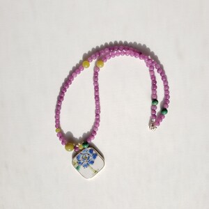 May include: A purple and green beaded necklace with a square pendant featuring a painted floral design.