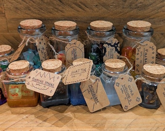 Puede incluir: Una colección de 14 pequeñas botellas de vidrio con tapas de corcho, cada una llena de dados coloridos y etiquetadas con nombres y niveles escritos a mano. Las etiquetas incluyen "Flaming Sphere", "Shatter", "Moonbeam", "Cure Wounds" y "Thunderwave".