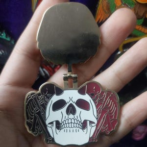 Puede incluir: Pin de metal dorado con un diseño de calavera. La calavera es blanca con detalles rojos y tiene cuernos. La calavera está rodeada por dos esqueletos con detalles rojos. El pin está unido a una cadena de metal dorado.