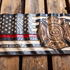 May include: A carved wooden American flag with a wavy texture, featuring a thin red horizontal stripe, a dark stained canton with stars, and a large central fire department emblem. The text reads JOHN RAYMER and CITY OF EAST PEORIA FIRE DEPT.