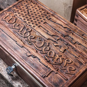 May include: A wooden box with a carved American flag design and the words "We the People." The box is brown with a lock and key. The design includes carved rifles and stars.