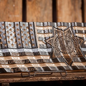 May include: A wooden American flag with a black and white wood grain pattern. The flag has a carved star in the center with the words "Department of Corrections" and "Seal of the State of Ohio" around the star. The flag also has the name "Joseph Jennings", "Deputy Commander of Intelligence", and "30 Years of Service" carved into the wood.