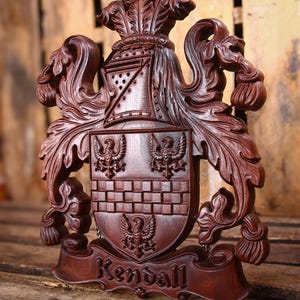 May include: A dark brown carved wooden coat of arms featuring a central shield with three eagle figures and a checkered pattern. Above the shield is a helmet and ornate foliage. A banner at the bottom displays the name Kendall in relief.