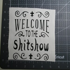 May include: A rectangular piece of light-colored fabric featuring black printed text that reads WELCOME TO THE SHITSHOW, accented with decorative swirls and fleur-de-lis symbols, positioned on a dark cutting mat with visible inch markings.