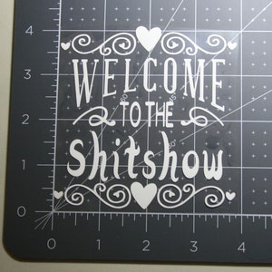 May include: A white vinyl decal featuring the text "WELCOME TO THE SHITSHOW" in a decorative font, accented with swirling flourishes and heart shapes. The decal is placed on a dark gray cutting mat with a visible grid marked in inches.