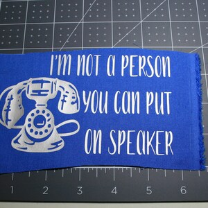 Puede incluir: Una pieza rectangular de tela azul con un gráfico blanco de un teléfono de disco vintage y el texto "I'M NOT A PERSON YOU CAN PUT ON SPEAKER" sobre una base de corte negra con líneas de cuadrícula blancas que miden hasta 20 cm.