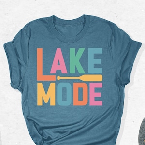 May include: A teal blue t-shirt with the text "LAKE MODE" in colorful block letters. A yellow paddle is included in the design. The text "SPECTRUM APPAREL" is in a white box in the upper right corner of the image.