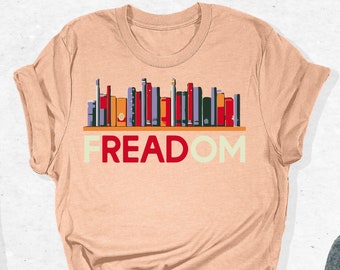 May include: A peach coloured t-shirt with a graphic of a bookshelf filled with colourful books. The text "FREEDOM" is printed below the bookshelf.