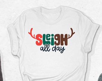 May include: White t-shirt with the text "SLEIGH all day" in red, green, brown, and blue. The shirt is paired with gray jeans and gray sneakers with brown laces. The shirt has a reindeer antler design.