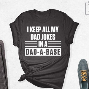 May include: A dark gray t-shirt with white text that reads "I keep all my dad jokes in a dad-a-base". The shirt is folded and tied in a knot. A pair of dark gray jeans and a pair of blue shoes are also pictured.