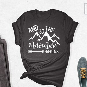 May include: A dark gray t-shirt with a white graphic design that says "And so the adventure begins." with a mountain range and an arrow pointing to the right.