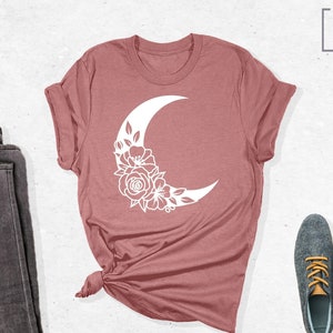May include: A mauve t-shirt with a white crescent moon design featuring floral details. The t-shirt is folded and displayed on a white background with a pair of dark grey jeans and a pair of grey shoes with yellow laces.