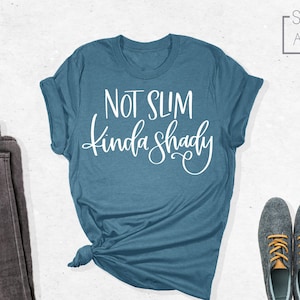 May include: A blue t-shirt with the text "Not Slim Kinda Shady" in white lettering. The shirt is folded and laid flat on a white surface.