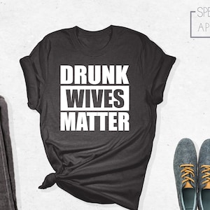 May include: A dark gray t-shirt with the text "DRUNK WIVES MATTER" printed in white.