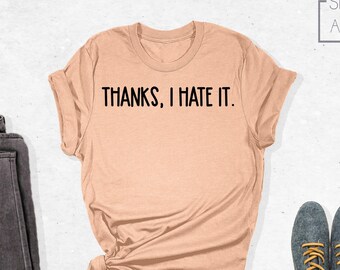 May include: A peach-colored t-shirt with the text "THANKS, I HATE IT." printed in black. The shirt is folded and laid on a white surface with a pair of dark gray jeans and two gray shoes with yellow laces.